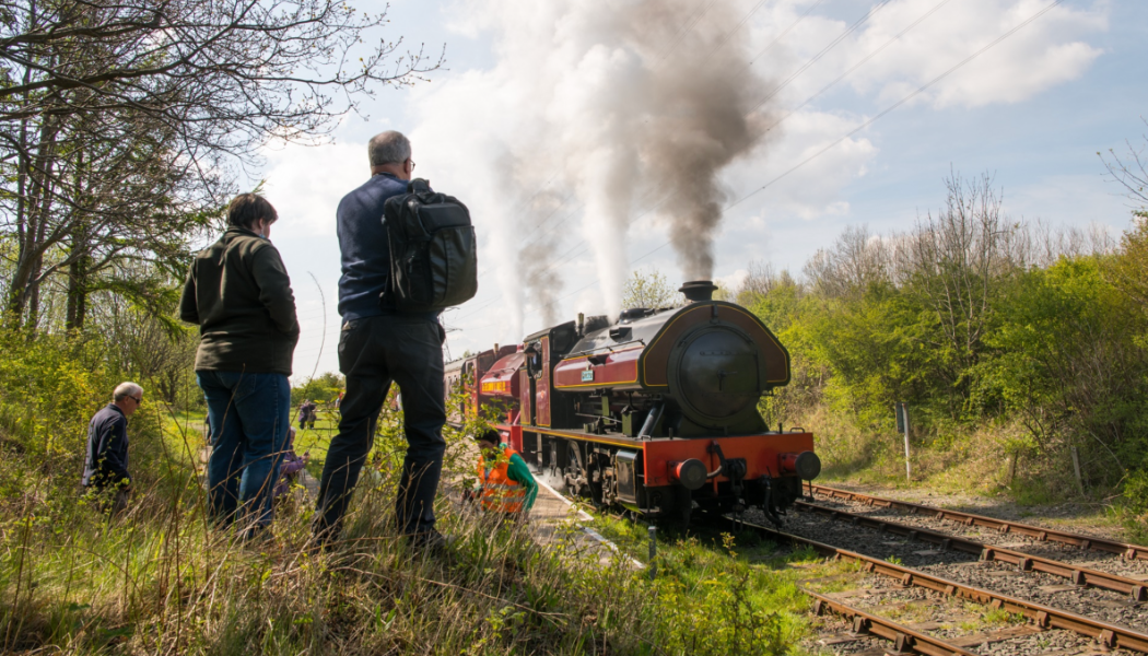 A steam engine travels along tracks as onlookers watch from an embankment